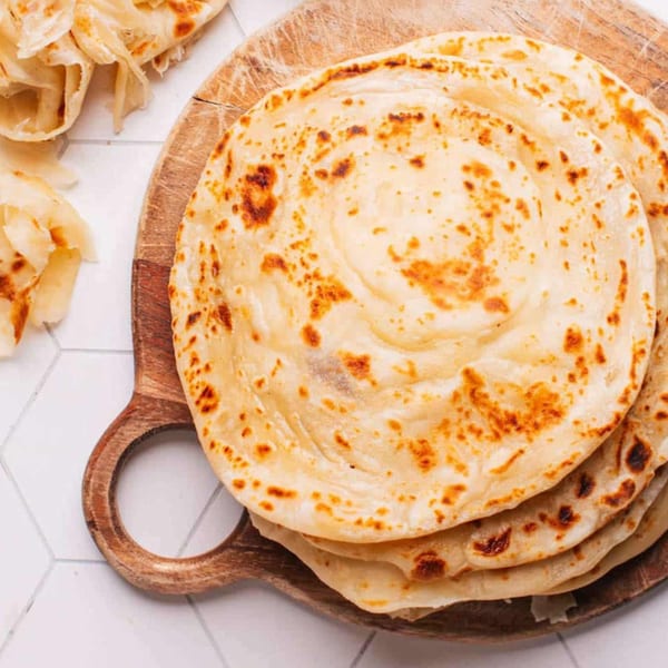 Stack of golden-brown paratha flatbreads on a wooden board with more flatbreads in the background.