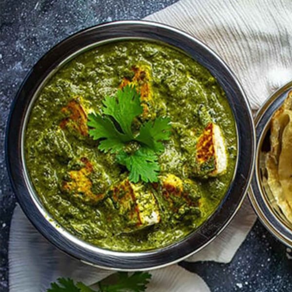 A silver bowl of vibrant green palak paneer with paneer cubes and cilantro garnish.